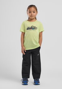 T-shirt vert clair avec un graphique de montagne et le texte "Nomades de la Nature", associé à un pantalon cargo noir et des chaussures bleues. Fond uni.