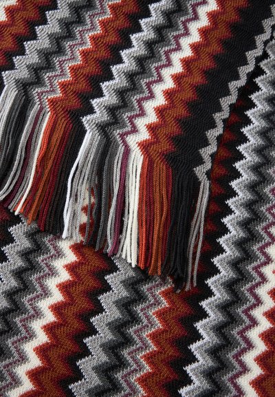 Tissu tricoté avec motif en zigzag noir, blanc, gris, bordeaux et orange brûlé, avec des franges multicolores sur le bord.