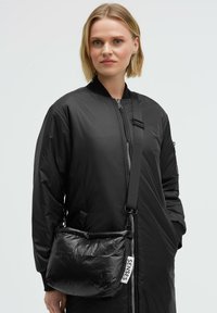 Schwarze Stepp-Tasche mit glänzender Textur, ausgestattet mit einem Schulterriemen und einem weißen Etikett mit der Aufschrift "SENSES". Kombiniert mit einer schwarzen Bomberjacke.