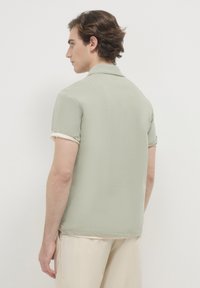 Polo shirt verde pallido con maniche arrotolate di colore chiaro, texture morbida e vestibilità rilassata, vista da dietro.