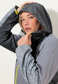 Veste imperméable grise avec une manche à motif floral, capuche réglable et accents jaune fluo. Texture lisse, design ajusté.