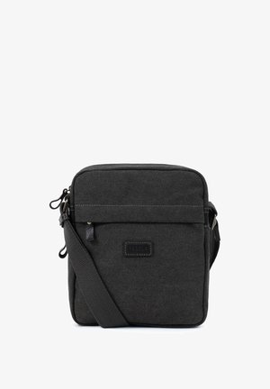 Petit sac bandoulière en tissu noir avec poche avant zippée, sangle réglable et patch logo Fagola visible centré sur le devant.