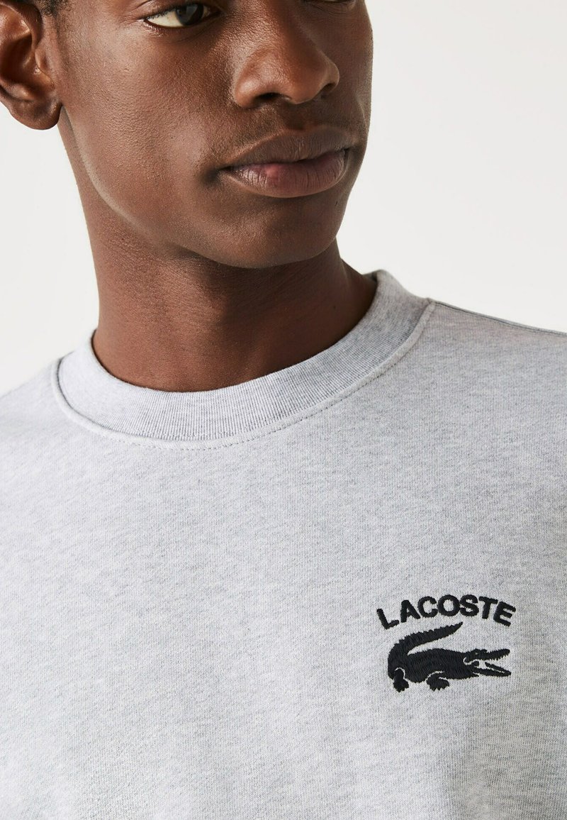 Sweatshirt gris en coton, avec un col rond et des poignets côtelés. Affiche un texte "Lacoste" brodé en noir et un logo de crocodile.