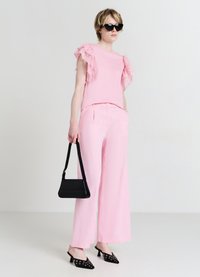 Haut rose à manches volantes, pantalon large rose, sac à main noir et ballerines noires à bouts pointus avec œillets décoratifs. Lunettes de soleil portées.