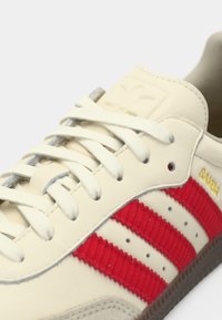 Κοντινό πλάνο μπεζ αθλητικού παπουτσιού Adidas Samba με κόκκινες ριγές με υφή, λευκά κορδόνια και ορατή σόλα από καουτσούκ.