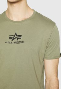 Oliven grøn bomull t-skjorte med rund hals og korte ermer, med et svart trykt logo "ALPHA INDUSTRIES" og et stilisert emblem.