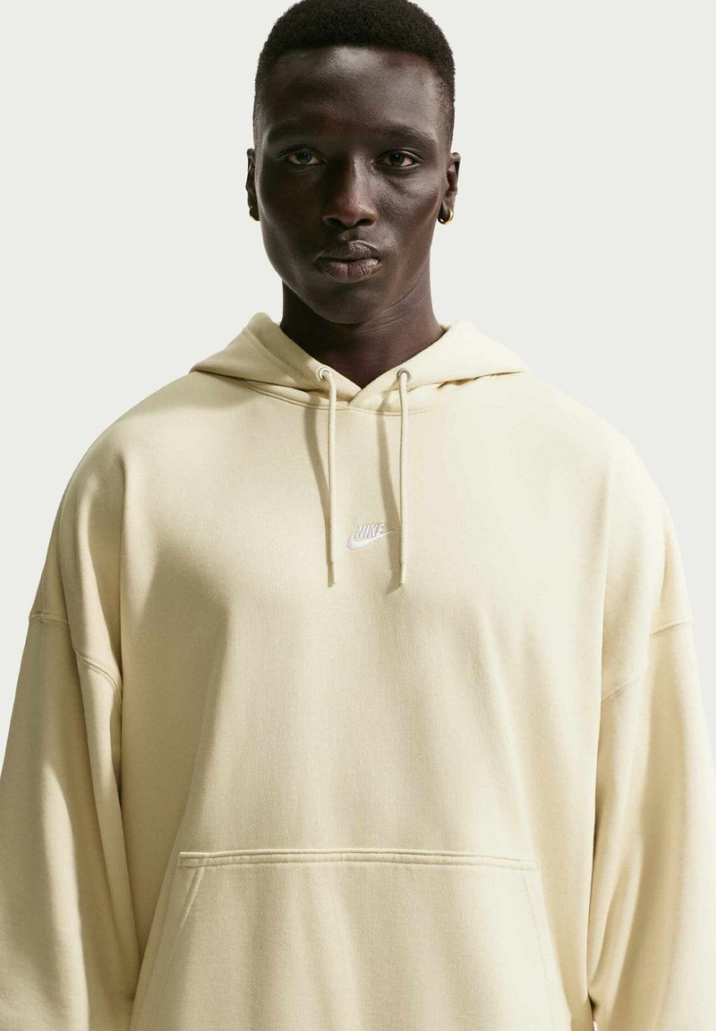 Personne portant un hoodie Nike beige clair avec une poche à l'avant, regardant droit devant sur un fond uni clair.