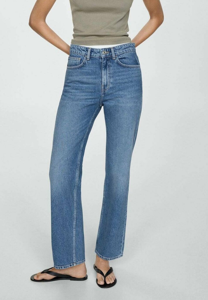 Femme portant un jean bleu taille haute à jambe droite, un haut sans manches vert olive et des tongs noires, debout, les mains sur les hanches.