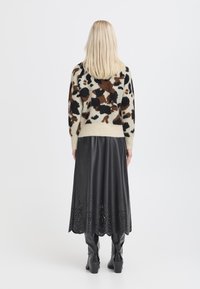 Maglione lavorato a maglia con sfondo beige e macchie nere/marroni, abbinato a una gonna in pelle nera con dettagli tagliati al laser lungo l'orlo.