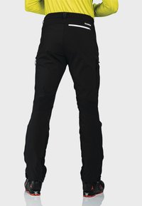 Schwarze Outdoor-Hosen mit taillierter Passform, ausgestattet mit seitlichen und hinteren Reißverschlusstaschen sowie verstellbaren Knöchelbündchen, getragen mit Skischuhen.