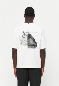 Hvid bomulds t-shirt med en sort-hvid grafik på ryggen, der inkluderer en illustration af en skorpion og topografiske kortdetaljer.