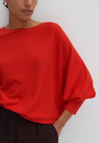 Pull rouge surdimensionné avec des manches chauve-souris, texture tricot douce, grand col et détails de couture, accompagné de pantalons foncés.