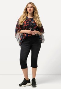 Top negro transparente con patrón abstracto colorido, combinado con jeans capri negros y zapatillas deportivas negras y gruesas. El cabello es largo y ondulado.