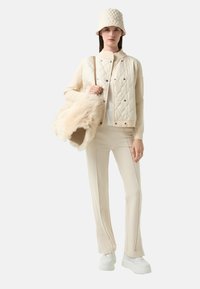 Kurtka w kolorze kremowym z pikowanym wzorem i zatrzaskami, dopasowana czapka typu bucket, biała bluzka, jasnobeżowe dopasowane spodnie oraz białe sneakersy na platformie.