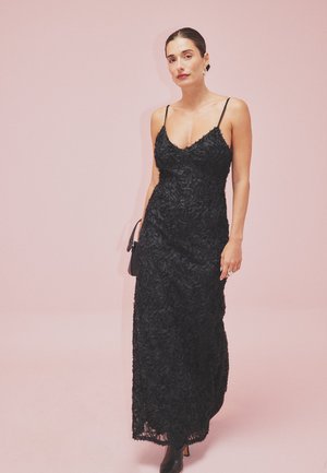 Abito maxi in pizzo nero con motivo floreale, silhouette aderente e spalline sottili. Il modello tiene una piccola borsa a tracolla nera su uno sfondo rosa.