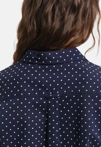 Navy blue shirt featuring a polka dot pattern in white. It has a collar and a smooth, textured fabric with a back yoke detail.