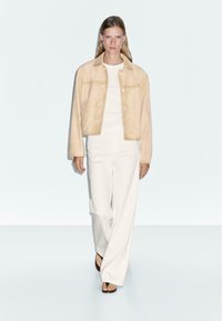 Beige suède jack met knoopsluiting, gecombineerd met witte wijde broeken en zwarte sandalen. Model heeft lang haar en eenvoudige accessoires.