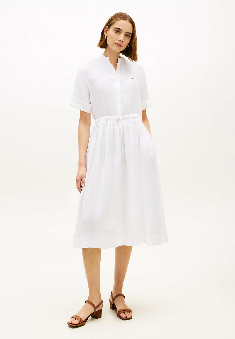 Femme debout portant une robe midi blanche à manches courtes boutonnée avec taille à cordon et sandales à talons marron sur un fond clair.