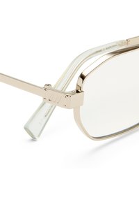 Ορθογώνια γυαλιά με χρυσό μεταλλικό πλαίσιο, διάφανα φακούς και διάφανα άκρα στα βραχίονες, που ακουμπούν σε λευκή επιφάνεια.