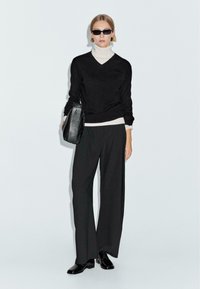 Maglione nero con scollo a V sopra un dolcevita bianco, abbinato a pantaloni neri a gamba larga. Una borsa nera e scarpe nere lucide completano il look.