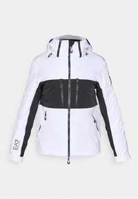 Λευκή και μαύρη σακάκι σκι με κουκούλα, που διαθέτει τσέπες με φερμουάρ, σχεδίαση χρωματικών μπλοκ αντίθεσης και υφασμάτινες διακοσμήσεις στους ώμους.
