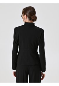 Zwarte op maat gemaakte blazer met een hoge kraag, een getailleerde vorm en lange mouwen. Heeft een getailleerde taille en witte hemdmanchetten zichtbaar bij de polsen.