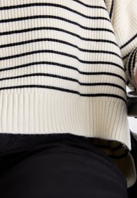 Maglione crema con strisce orizzontali nere, orlo a coste e tessuto textured, abbinato a pantaloni scuri nella parte inferiore.