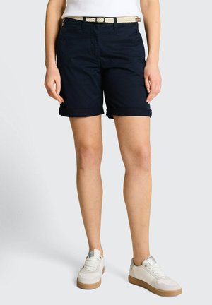 Kobieta w granatowych szortach z podwiniętymi nogawkami, białym plecionym paskiem, białych trampkach na gumowej podeszwie i białej, wsuniętej bluzce, stojąca na jednolitym tle.