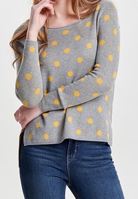 Pull en maille grise à pois jaunes vifs, avec un col rond et des manches longues, assorti à un jean bleu.