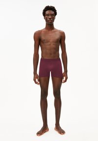 Burgunderfarbene Boxershorts mit anliegendem Design, hergestellt aus weichem Stoff, mit einem breiten Bund und Branding. Das Modell steht barfuß in neutraler Pose.