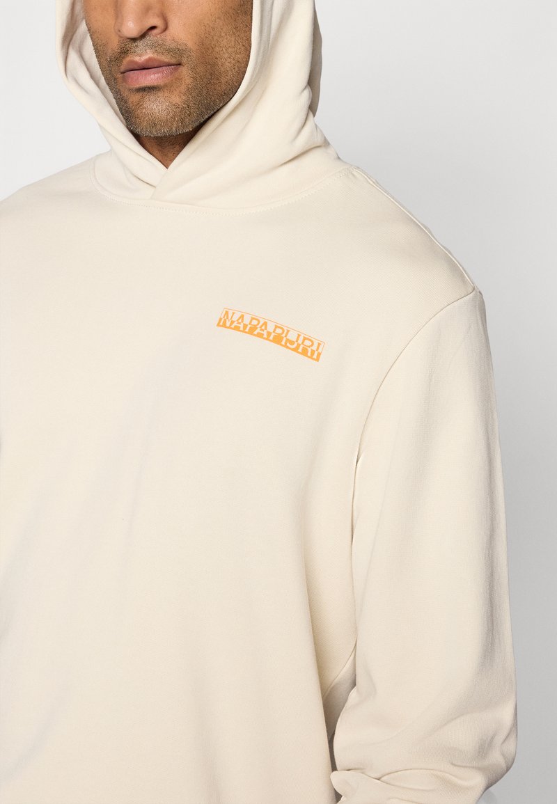 Beige Kapuzensweatshirt mit weicher Textur, auf der Brust mit einem orangefarbenen Logo-Patch. Lockerer Schnitt mit einem einfachen, cleanen Design.
