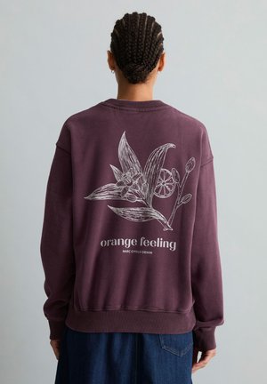 Person mit geflochtenem Haar, die ein lila Sweatshirt mit weißem Blumen- und Orangenscheibengrafik sowie dem Text "orange feeling" auf dem Rücken trägt.