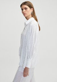 Witte blouse met verticale strepen, versierd met kleine zilveren studs. Lange mouwen met manchetten, op maat gemaakte uitstraling. Soepele textuur.