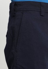 Gros plan sur la poche et la ceinture bleu marine d'un pantalon avec passants pour ceinture, porté avec une chemise bleue rentrée.