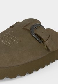 Sabot en daim marron avec boucle argentée et motif cousu sur le dessus, doté d'une semelle épaisse rainurée pour une bonne adhérence.