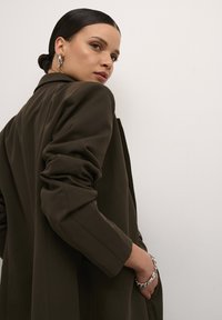 Olivenfarvet skræddersyet jakke med strukturerede skuldre og lange ærmer, med en lomme. En sølv kædearmbånd fremhæver håndleddet.