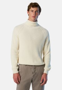 Maglione a collo alto a costine di colore crema, con maniche lunghe e silhouette aderente, abbinato a pantaloni marrone chiaro. Dettaglio del tessuto testurizzato visibile.