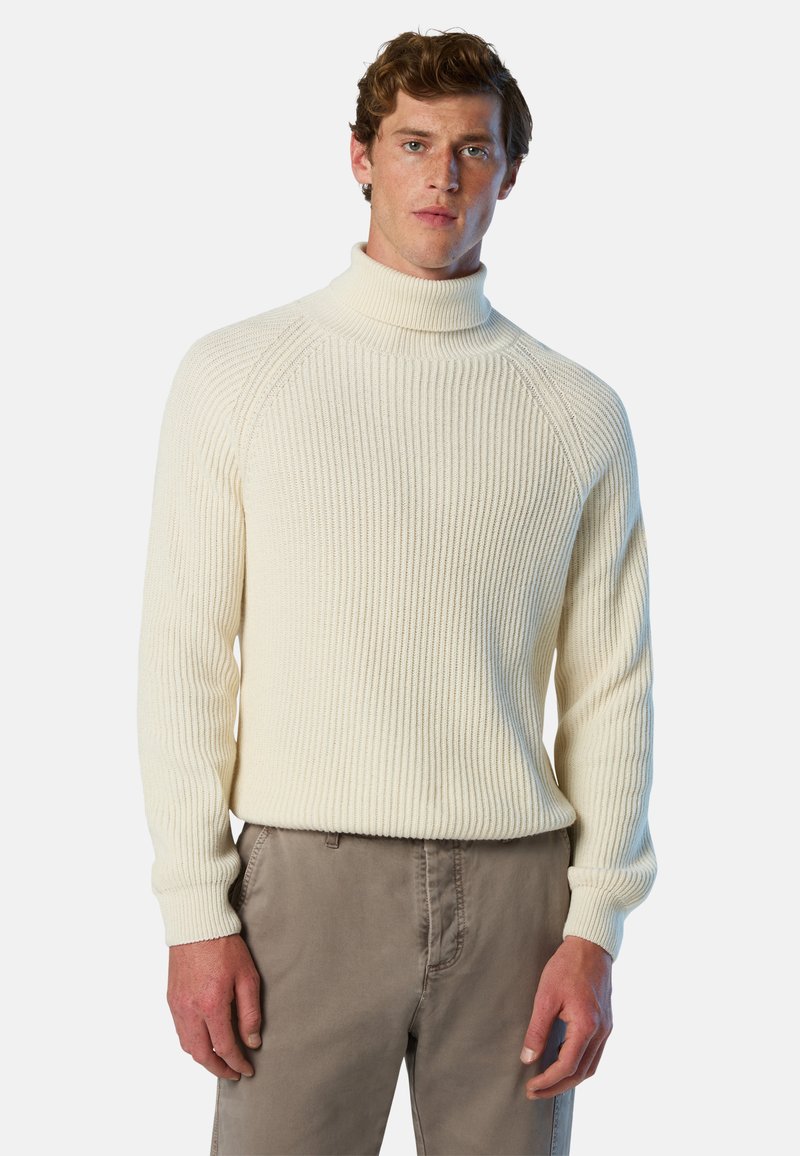 Maglione a collo alto a costine di colore crema, con maniche lunghe e silhouette aderente, abbinato a pantaloni marrone chiaro. Dettaglio del tessuto testurizzato visibile.