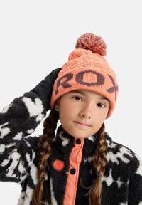 Knitted orange beanie with a textured pattern and a top pom-pom, featuring bold "ROXY" lettering in dark brown. Worn with a black and white fleece jacket.