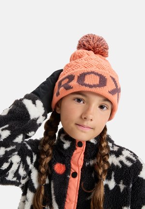 Bonnet orange tricoté avec un motif texturé et un pompon sur le dessus, arborant un lettrage audacieux "ROXY" en brun foncé. Porté avec une veste polaire noire et blanche.