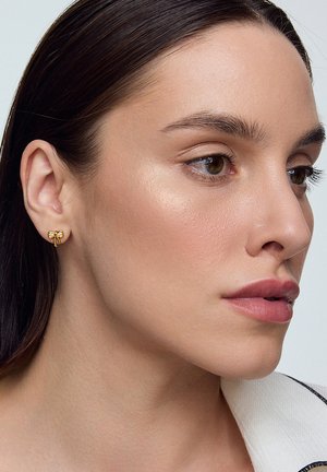 Złote kolczyki w kształcie kokardy z małymi akcentami z cyrkonii. Tło jest neutralne, co podkreśla szczegóły dodatku.