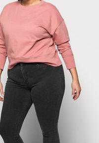 Różowy bawełniany sweatshirt z okrągłym dekoltem i opadłymi ramionami, zestawiony z czarnymi wąskimi dżinsami o klasycznym pasie i kieszeniach.
