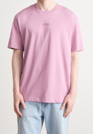 Osoba nosząca gładki, jasnofioletowy t-shirt z krótkim rękawem z napisem "BOSS" na piersi oraz jasnoniebieskie dżinsy na jednolitym tle.