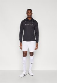 Svart långärmad tröja i jerseymaterial med krage och grå logotyp, kombinerad med vita shorts och vita knälånga strumpor med svart logotyp.