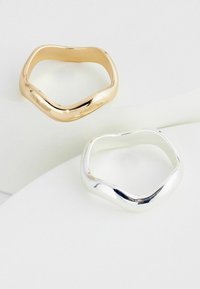 Zwei Ringe präsentiert: ein eleganter goldener Ring und ein silberner Ring, beide mit einer wellenartigen Form und einer polierten, reflektierenden Oberfläche auf einem hellen Hintergrund.