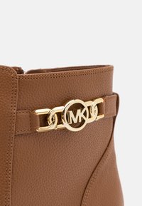 Botte en cuir marron texturé avec un détail de chaîne dorée au logo MK près du bord supérieur sur un fond uni.