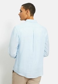 Camicia in lino azzurro chiaro con plissettatura sul dietro, morbida al tatto, maniche lunghe arrotolate fino al gomito e colletto.