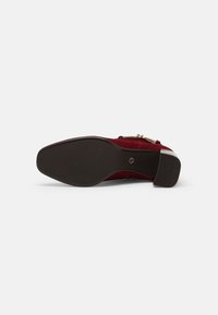 Chaussure en daim rouge à semelle plate vue par la semelle, avec un petit talon bloc, une texture antidérapante et une boucle dorée sur le dessus.