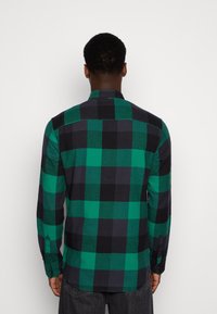 Hombre vestido con una camisa de manga larga a cuadros verdes y negros, de espaldas, de pie frente a un fondo blanco liso.