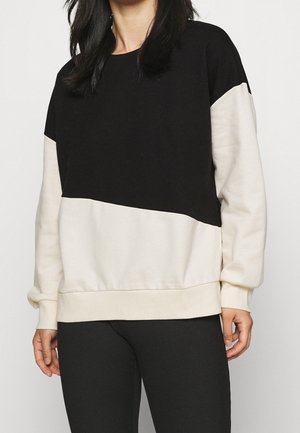 Vrouw die een zwart-wit sweatshirt met lange mouwen draagt, gecombineerd met zwarte geribbelde legging, staand tegen een effen achtergrond.